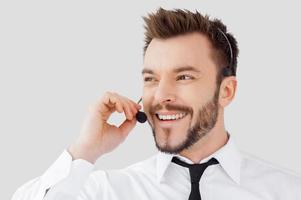 Cheerful customer service representative. Handsome young man in formalwear and headset looking away and smiling while standing against grey background photo