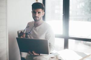 Always ready to help you. Confident young man carrying laptop and eyeglasses while leaning at the desk in office and looking at camera photo