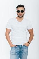Feeling confident in my skin. Handsome young Indian man in sunglasses holding hands in pockets and looking at camera while standing against white background photo