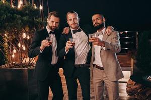 Three cheerful men in suits holding whiskey glasses and looking at camera while spending time on party photo