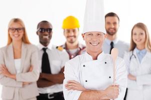 When I grow up I will be a chef. Confident male chef in uniform keeping arms crossed and smiling while group of people in different professions standing in the background photo