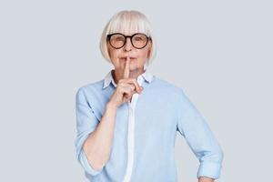 guarde silencio. hermosa anciana manteniendo el dedo en los labios mientras está de pie contra el fondo gris foto