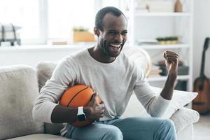 Feeling happy. Handsome young African man holding a ball and laughing while sitting on the sofa at home photo