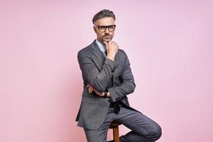 Confident mature man in formalwear holding hand on chin while sitting against pink background photo