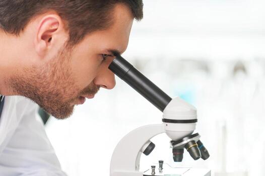 Concentrated on cure. Side view of young scientist in white uniform using microscope while sitting at his working place photo