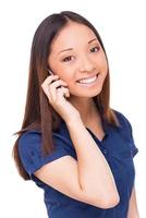 Beauty on the phone. Cheerful young Asian woman talking on the mobile phone and looking at camera while standing isolated on white photo