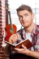 creativo dentro de su alma. un joven apuesto escribiendo algo en un bloc de notas y tocándose la barbilla con un bolígrafo mientras la guitarra acústica está en el fondo foto