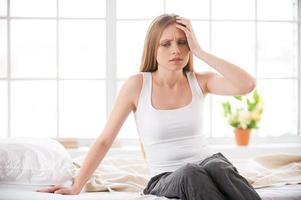 sintiendo dolor de cabeza. mujer joven frustrada tocando la cabeza con la mano y expresando negatividad mientras se sienta en la cama en su apartamento foto