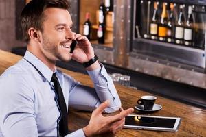 buena charla con un amigo. vista lateral de un joven feliz con camisa y corbata hablando por teléfono móvil y gesticulando mientras se sienta en el mostrador del bar foto