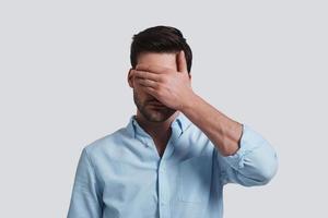 See nothing.  Handsome young man covering eyes with hands while standing grey background photo