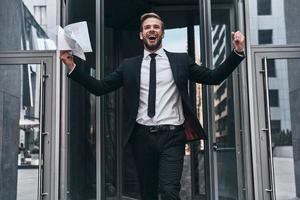 apuesto joven hombre de negocios con traje completo sonriendo y sosteniendo un documento mientras camina al aire libre foto