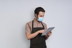 Handsome young man in apron wearing protective face and using digital tablet while standing against gray background photo