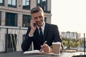 Cheerful mature businessman talking on mobile phone and making notes while sitting in cafe outdoors photo