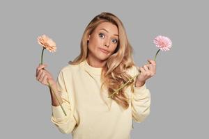 Which is more beautiful  Attractive young woman holding two flowers and looking at camera while standing against grey background photo