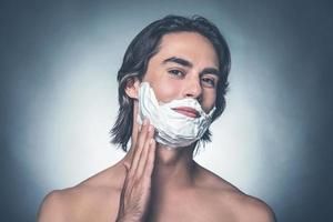 Comfortable shaving. Handsome young shirtless man spreading shaving cream over face and looking at camera while standing against grey background photo