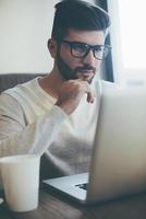 Thinking about solution. Thoughtful young man in glasses working on laptop and holding hand on chin while sitting in office or cafe photo