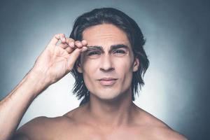 This is painful. Portrait of young shirtless man tweezing eyebrows and expressing negativity while standing against grey background photo