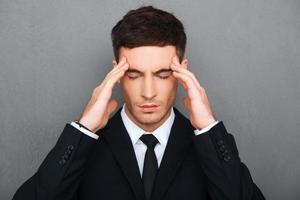 tratando de concentrarme. joven empresario deprimido tocándose la frente y manteniendo los ojos cerrados mientras se enfrenta a un fondo gris foto