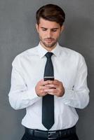 Sending business message. Handsome young man in shirt and tie holing mobile phone and looking at it while standing against grey background photo