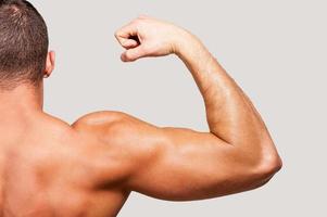 Showing his perfect bicep. Rear view of young muscular man showing his bicep on one hand while standing against grey background photo