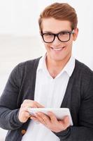 Cheerful IT guy. Handsome young man working on digital tablet and smiling while standing indoors photo