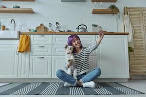 Playful woman carrying little dog and making selfie while sitting on the floor at the kitchen photo