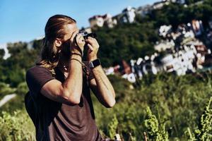 Great shot. Young man in casual clothing photographing the view while standing on the hill outdoors photo