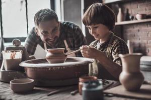 Time to create something great. Little boy drawing on ceramic pot at the pottery class while man in apron standing close to him photo