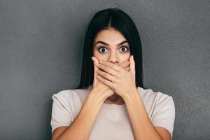 Trying to stop gossiping. Surprised young woman in casual wear covering mouth with hands and looking at camera while standing against grey background photo