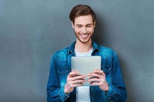 Examining his new gadget. Happy young man holding digital tablet and smiling while standing against grey background photo