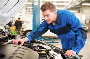 Man repairing car. Concentrated young man in uniform repairing car while standing in workshop photo