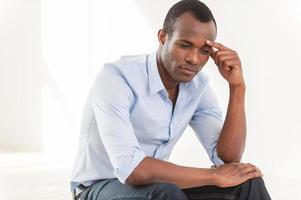 Thinking about... Thoughtful young African man in blue shirt touching forehead with hand and looking away photo