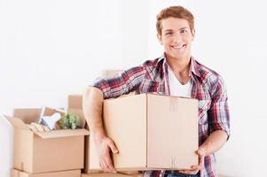 Moving to a new apartment. Cheerful young man holding a cardboard box and smiling while other carton boxes laying on background photo