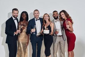 Group of happy beautiful people in formalwear holding birthday cake and champagne flutes photo