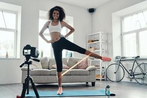 Full length of attractive young African woman exercising using strap and smiling while making social media video photo