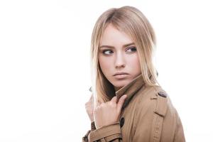 Beauty in coat. Attractive young woman in coat adjusting her collar and looking away while standing against white background photo