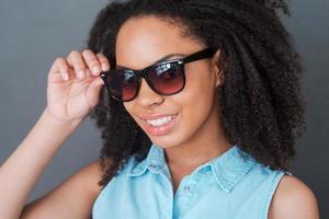 Beauty in style. Portrait of attractive young African woman adjusting her sunglasses and looking at camera with smile while standing against grey background photo