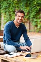 rostro de la satisfacción laboral. un joven sonriente que trabaja en una laptop y mira la cámara mientras se sienta en la mesa de madera al aire libre foto