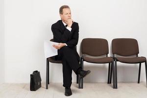 Job candidate. Bored mature man in formalwear holding paper and keeping legs crossed at knee while sitting at the chair in waiting room photo