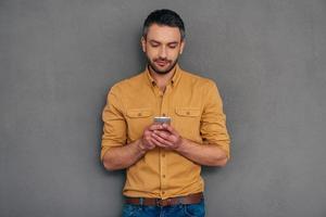 Quick message to friend. Confident mature man holding smart phone and looking at it while standing against grey background photo