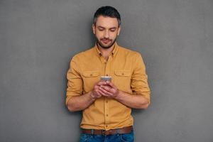 Quick message to friend. Confident mature man holding smart phone and looking at it while standing against grey background photo