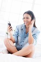 Starting day with her favorite music. Happy young woman in headphones listening to MP3 player and smiling while sitting in bed photo