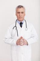 Taking care of your health. Senior grey hair doctor in uniform looking at camera and keeping hands clasped while standing isolated on white photo