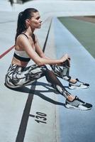 Towards a healthier lifestyle. Beautiful young woman in sports clothing relaxing while sitting on the running track outdoors photo
