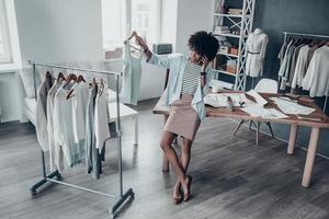 Full length of attractive young African woman talking on smart phone and holding a clothes rack while leaning on the desk in workshop photo