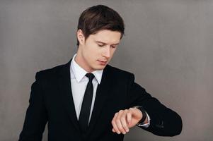 comprobando la hora. joven confiado en ropa formal mirando su reloj de pulsera mientras está de pie contra el fondo gris foto