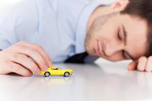 soñando con un coche deportivo. un joven alegre con camisa y corbata jugando con un vehículo de juguete mientras se sienta en su lugar de trabajo foto