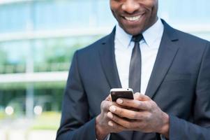 Typing business message. Cropped image of cheerful young African man in formalwear holding mobile phone and smiling while standing outdoors photo