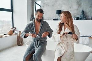 amorosa pareja joven en albornoces sonriendo y limpiando los dientes mientras hace la rutina matutina foto