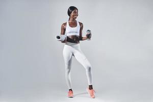 Happy young African woman in sports clothing holding bottle with water and exercise mat photo